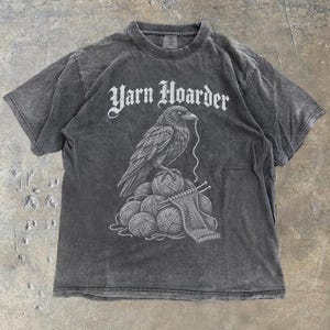 May include: A charcoal gray short-sleeve t-shirt featuring a graphic print of a raven perched on a pile of yarn balls with knitting needles. The text "Yarn Hoarder" is printed in a white gothic-style font above the raven illustration.