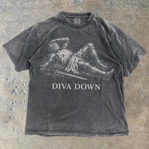 May include: A charcoal gray short-sleeve t-shirt with a faded wash. The front features a graphic illustration of a knight in armor lying on the ground with a sword nearby. Below the graphic, the text DIVA DOWN is printed in a white serif font.