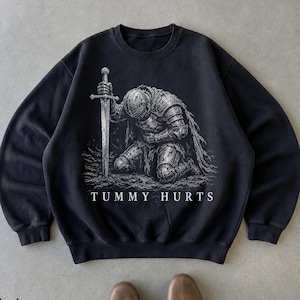 May include: A black crewneck sweatshirt featuring a white graphic print of a knight in full armor kneeling and holding a sword. Below the illustration, the text "TUMMY HURTS" is printed in a clean, white, capitalized sans-serif font.