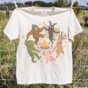 Puede incluir: Una camiseta de manga corta color marfil con un estampado gráfico de un sasquatch marrón, una criatura del pantano verde, un ajolote rosa, una polilla y un jackalope bailando en círculo alrededor de una fogata, rodeados de setas y lunas.
