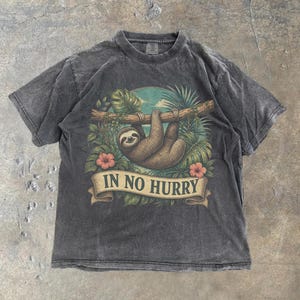 May include: A charcoal grey short-sleeve t-shirt featuring a graphic of a brown sloth hanging from a tree branch surrounded by green tropical leaves and pink hibiscus flowers. Below the sloth is a tan banner with the text IN NO HURRY.