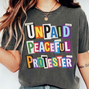 以下が含まれることがあります： 「UNPAID PEACEFUL PROTESTER」の文字がカラフルな切り抜き文字でプリントされた、濃いグレーのTシャツ。クルーネックで半袖です。モデルはブルージーンズを着用しています。