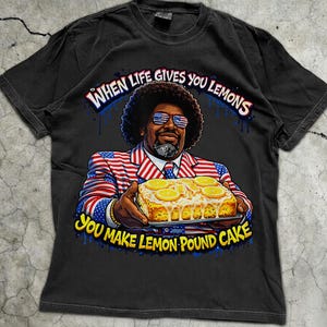Puede incluir: Camiseta negra con un gráfico colorido de un hombre con un traje patriótico sosteniendo un bizcocho de limón. El texto dice: "Cuando la vida te da limones, haz bizcocho de limón."