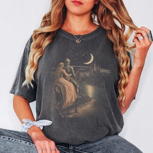 May include: A charcoal grey short-sleeve t-shirt featuring a graphic print of a knight in armour and a person in a long dress riding a horse towards a castle under a crescent moon and starry night sky. The shirt has a crew neckline and a relaxed fit.