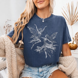 May include: A dark blue short-sleeve t-shirt featuring a white graphic print of two fairies dancing amidst scattered stars. The shirt has a crew neckline and a relaxed fit, paired with light blue denim shorts and a textured beige cardigan.