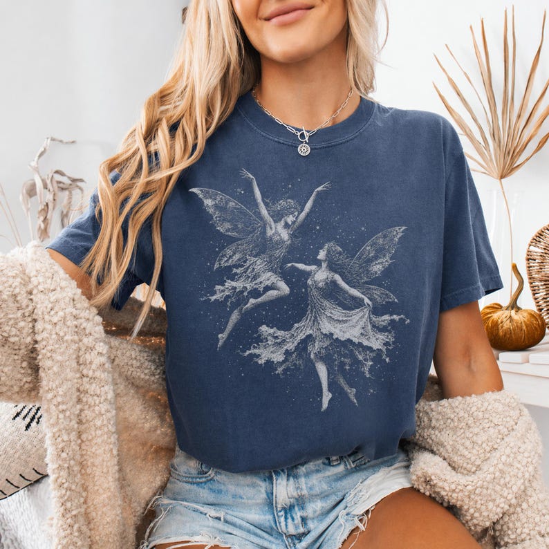 May include: A dark blue short-sleeve t-shirt featuring a white graphic print of two fairies dancing amidst scattered stars. The shirt has a crew neckline and a relaxed fit, paired with light blue denim shorts and a textured beige cardigan.