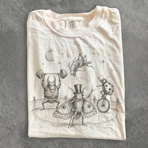 Puede incluir: Una camiseta color crema con una ilustración en tinta negra de insectos de circo. Un escarabajo levanta una pesa, una polilla lleva un sombrero de copa, una mariquita monta un monociclo y un saltamontes salta bajo una luna creciente y estrellas.