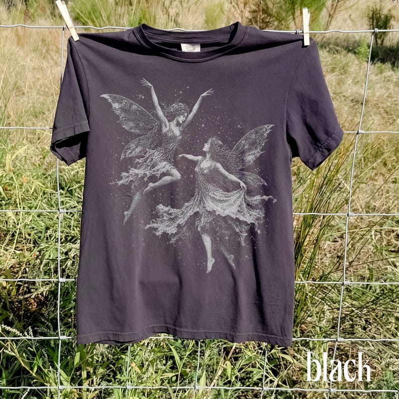 May include: A charcoal black short-sleeve t-shirt featuring a white graphic print of two winged fairies dancing amidst a field of small stars. The design is centered on the chest, showcasing detailed wings and flowing garments on both figures.