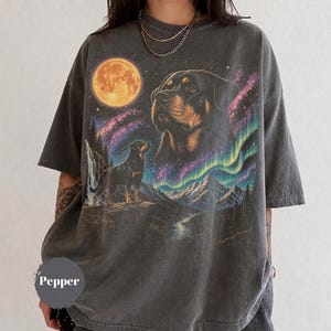 Könnte beinhalten: Ein anthrazitfarbenes Kurzarm-T-Shirt mit einem Grafikdruck eines Rottweilers, eines Vollmonds, einer Berglandschaft und farbenfrohen Nordlichtern. Das T-Shirt hat einen Rundhalsausschnitt und eine lockere, entspannte Passform mit verwaschenem Stoff.