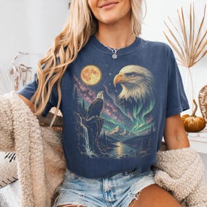 Peut inclure: Un t-shirt bleu fonc&eacute; &agrave; manches courtes avec une impression graphique repr&eacute;sentant un pygargue &agrave; t&ecirc;te blanche perch&eacute; sur une falaise, une grande t&ecirc;te d'aigle, une pleine lune, un ciel &eacute;toil&eacute;, une cascade, des montagnes et des aurores bor&eacute;ales.