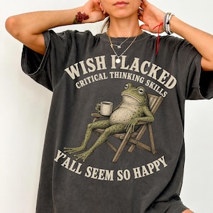 May include: A charcoal grey short-sleeve t-shirt featuring a graphic of a green frog sitting in a wooden chair holding a white mug. The text reads WISH I LACKED CRITICAL THINKING SKILLS Y'ALL SEEM SO HAPPY in a distressed off-white font.
