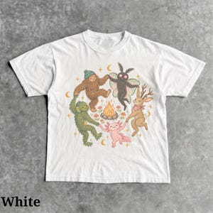 May include: A white short-sleeve t-shirt featuring a central graphic of a brown sasquatch, a green swamp creature, a pink axolotl, a black moth, and a brown jackalope holding hands in a circle around a small campfire with yellow crescent moons.