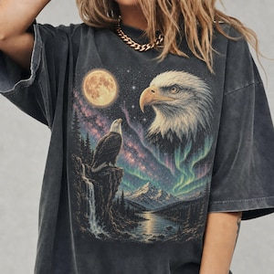 Peut inclure: Un t-shirt gris anthracite oversize avec une impression graphique repr&eacute;sentant un pygargue &agrave; t&ecirc;te blanche perch&eacute; sur une falaise pr&egrave;s d'une cascade, une grande t&ecirc;te d'aigle, une pleine lune, un ciel &eacute;toil&eacute; et des aurores bor&eacute;ales vertes.