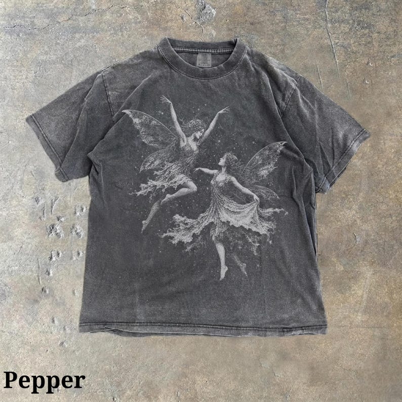 May include: A charcoal gray short-sleeve t-shirt featuring a white graphic print of two winged fairies dancing amidst a starry background. The fabric has a washed, faded appearance, and the word Pepper is printed in black at the bottom left.