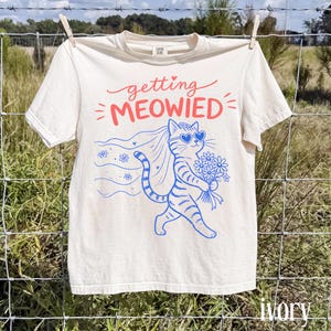 May include: An ivory short-sleeve t-shirt featuring a graphic of a cat wearing heart-shaped sunglasses and a veil, holding a bouquet of flowers. The text above reads "getting MEOWIED" in red, while the cat illustration is in blue.
