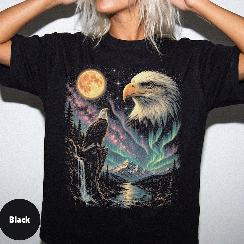 Peut inclure: Un t-shirt noir avec une impression graphique repr&eacute;sentant un pygargue &agrave; t&ecirc;te blanche perch&eacute; sur une falaise au-dessus d'une cascade, une grande t&ecirc;te d'aigle dans le ciel, une pleine lune, une galaxie &eacute;toil&eacute;e et des aurores bor&eacute;ales vertes.