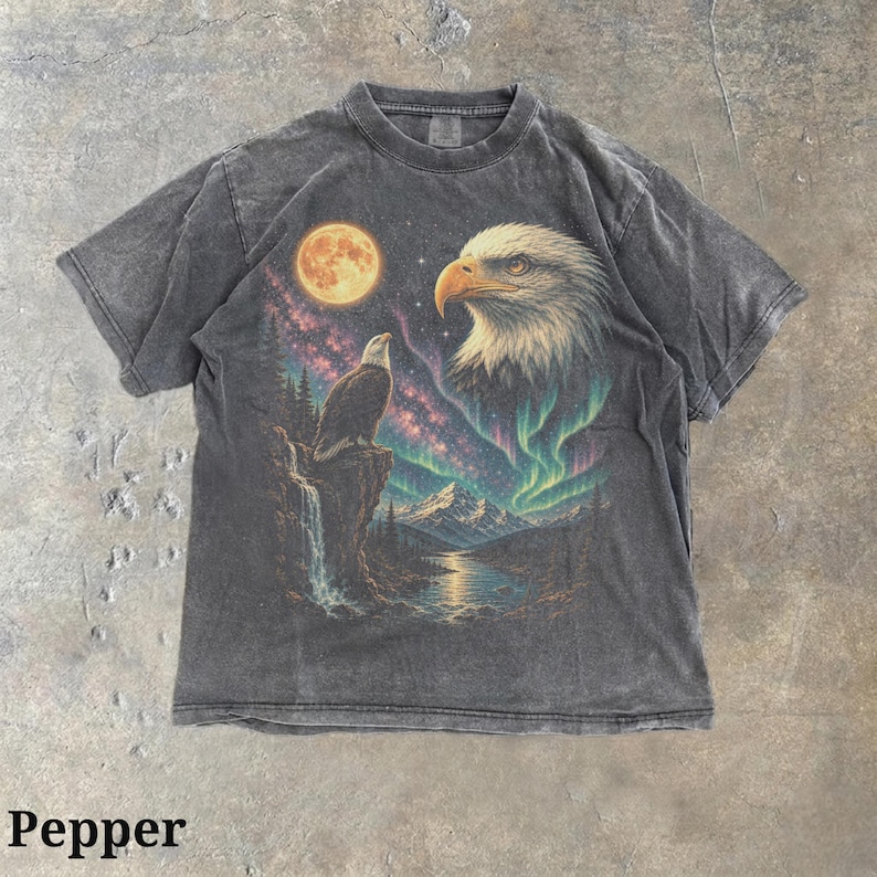 Peut inclure: Un t-shirt gris anthracite &agrave; manches courtes avec une impression graphique repr&eacute;sentant un pygargue &agrave; t&ecirc;te blanche perch&eacute; sur une falaise, une grande t&ecirc;te d'aigle, une pleine lune, un ciel &eacute;toil&eacute;, une cascade, une rivi&egrave;re et des aurores bor&eacute;ales.