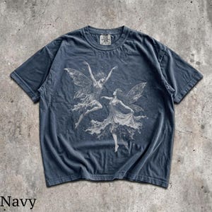 May include: A navy blue short-sleeve t-shirt featuring a white graphic print of two winged figures dancing. The shirt has a crew neckline and a visible brand tag at the inner collar. The fabric has a washed, faded appearance throughout.