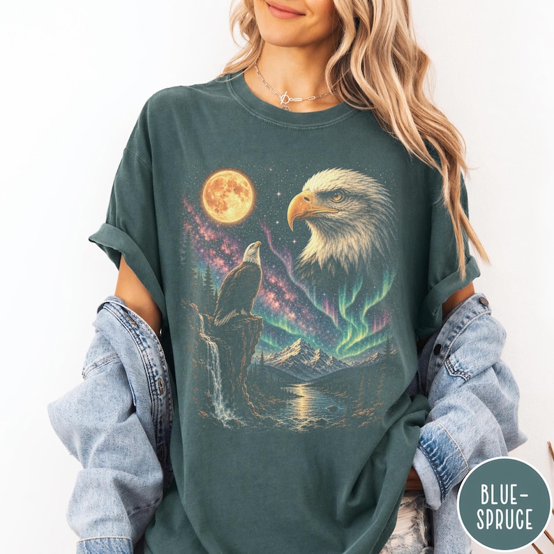 Peut inclure: Un t-shirt bleu sarcelle fonc&eacute; avec une impression graphique repr&eacute;sentant un pygargue &agrave; t&ecirc;te blanche perch&eacute; sur une falaise, une grande t&ecirc;te d'aigle, une pleine lune, une n&eacute;buleuse &eacute;toil&eacute;e et un paysage montagneux avec une rivi&egrave;re. Texte "BLUE-SPRUCE".