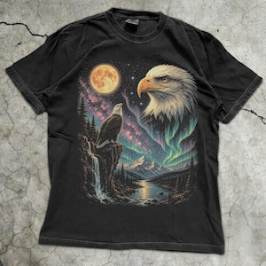Peut inclure: Un t-shirt noir &agrave; manches courtes avec une impression graphique repr&eacute;sentant un pygargue &agrave; t&ecirc;te blanche perch&eacute; sur une falaise au-dessus d'une cascade, une grande t&ecirc;te d'aigle, une pleine lune, un ciel &eacute;toil&eacute; et des aurores bor&eacute;ales vertes.