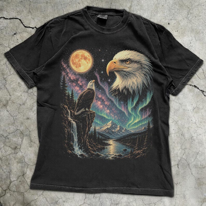Peut inclure: Un t-shirt noir &agrave; manches courtes avec une impression graphique repr&eacute;sentant un pygargue &agrave; t&ecirc;te blanche perch&eacute; sur une falaise au-dessus d'une cascade, une grande t&ecirc;te d'aigle, une pleine lune, un ciel &eacute;toil&eacute; et des aurores bor&eacute;ales vertes.