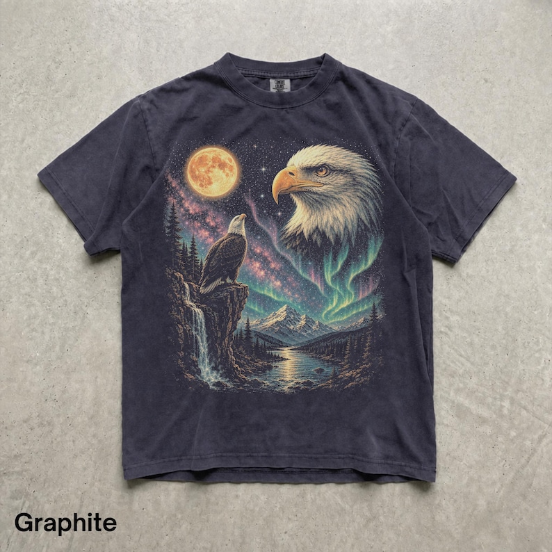 Peut inclure: Un t-shirt &agrave; manches courtes de couleur graphite avec une impression graphique repr&eacute;sentant un pygargue &agrave; t&ecirc;te blanche perch&eacute; sur une falaise surplombant une rivi&egrave;re, une grande lune, un ciel &eacute;toil&eacute; et un paysage montagneux avec une aurore bor&eacute;ale.