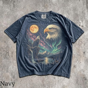 Peut inclure: Un t-shirt bleu marine avec une impression graphique repr&eacute;sentant un pygargue &agrave; t&ecirc;te blanche perch&eacute; sur une falaise, une grande t&ecirc;te d'aigle, une pleine lune, une cascade et des aurores bor&eacute;ales vertes au-dessus d'un paysage de montagne.