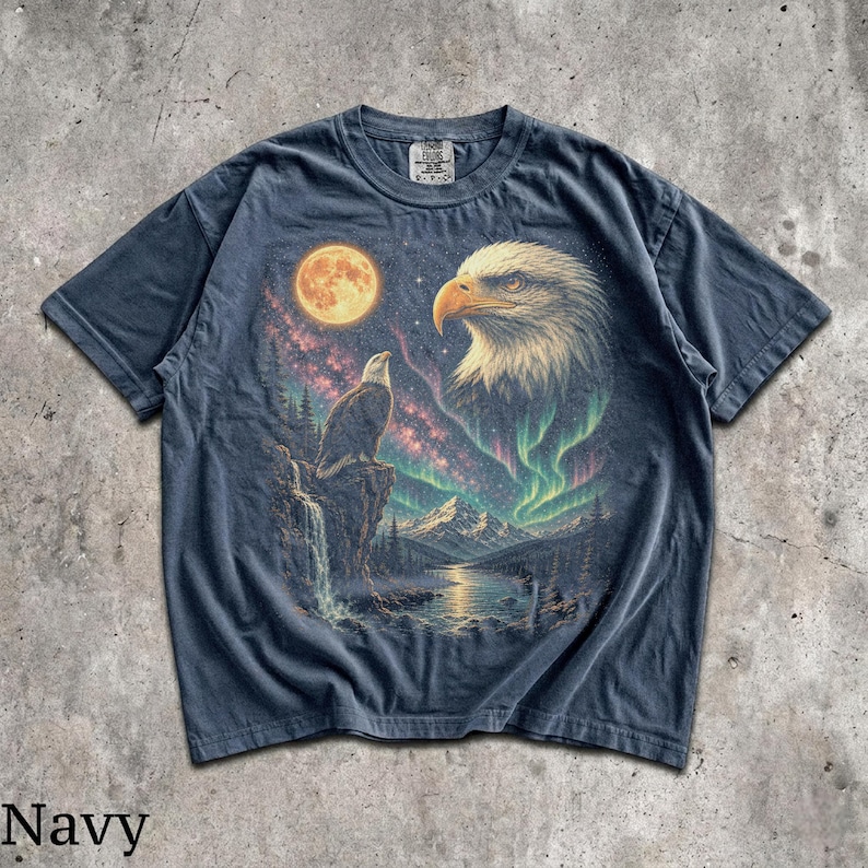 Peut inclure: Un t-shirt bleu marine avec une impression graphique repr&eacute;sentant un pygargue &agrave; t&ecirc;te blanche perch&eacute; sur une falaise, une grande t&ecirc;te d'aigle, une pleine lune, une cascade et des aurores bor&eacute;ales vertes au-dessus d'un paysage de montagne.