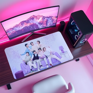 Puede incluir: Una configuración de escritorio de computadora con un monitor curvo, una computadora de escritorio negra y una alfombrilla de ratón rectangular. La alfombrilla muestra un grupo de personas en un sofá morado con confeti. El escritorio es marrón y la habitación está iluminada con luces rosas.