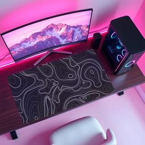 Könnte beinhalten: Ein schwarz-weißes Mauspad mit topografischem Design liegt auf einem Holzschreibtisch, neben einem gebogenen Computermonitor, der eine Berglandschaft zeigt. Ein schwarzer Computerturm mit bunten Lichtern steht ebenfalls auf dem Schreibtisch. Der Hintergrund ist rosa.