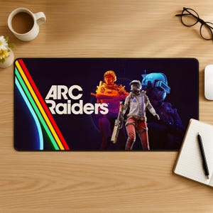 May include: A black rectangular mouse mat with the text "ARC Raiders" and a neon design. The mat features three figures in futuristic military attire. A coffee cup, glasses, and a notepad are on the wooden surface.