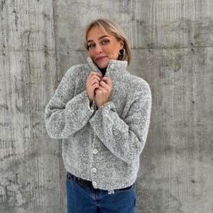 Può includere: Un cardigan in maglia screziata grigia e bianca con collo alto e chiusura con bottoni. Il maglione a vita corta è indossato con jeans blu e una cintura nera. I bottoni sono bianchi e il maglione ha le maniche lunghe.