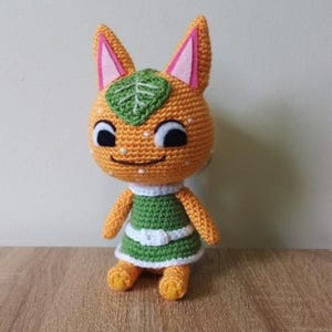 Op de afbeelding: Handgemaakte gehaakte oranje kattenpop. De pop heeft een groene en witte jurk, zwarte ogen en een lachend gezicht. De oren zijn oranje met roze binnenkanten. Een groen blad zit op het hoofd. Een knuffel.