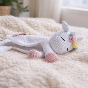 Può includere: Un giocattolo unicorno bianco all'uncinetto con zoccoli rosa e un copricapo floreale. L'unicorno è sdraiato su una coperta strutturata color crema. Il giocattolo ha gli occhi chiusi, suggerendo che stia dormendo.