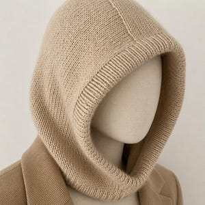May include: A beige knitted hood, displayed on a mannequin head, with a ribbed neckline and a seam running down the center. The hood is made of a soft, textured yarn and is paired with a tan blazer.