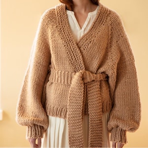 Puede incluir: Un cárdigan de punto grueso de color beige con un diseño envolvente y un cinturón a juego. El suéter presenta mangas largas y voluminosas y puños acanalados. El cárdigan se lleva sobre una camisa blanca con cuello y una falda blanca.