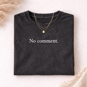 Könnte beinhalten: Dunkelgraues T-Shirt mit dem weißen Text "No comment.". Eine goldfarbene Halskette mit mehreren Reihen und einem kleinen Anhänger liegt auf dem Shirt. Das Shirt ist sauber auf einer hellen Oberfläche gefaltet.