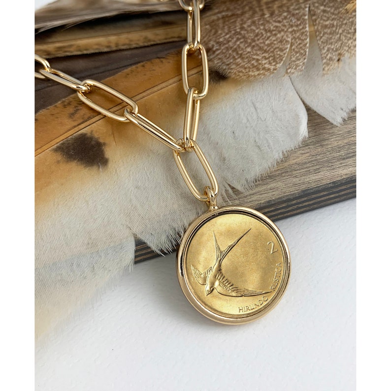 K&ouml;nnte beinhalten: Goldkette mit einem goldenen M&uuml;nzenanh&auml;nger, der ein Schwalbenmotiv und den Text "2 HIRL.DO" zeigt.