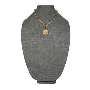 K&ouml;nnte beinhalten: Eine goldene Kettenkette mit einem gro&szlig;en runden Anh&auml;nger mit einem stilisierten Design. Die Kette wird auf einer grauen Stoffpuppe pr&auml;sentiert.