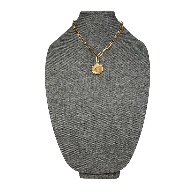K&ouml;nnte beinhalten: Eine goldene Kettenkette mit einem gro&szlig;en runden Anh&auml;nger mit einem stilisierten Design. Die Kette wird auf einer grauen Stoffpuppe pr&auml;sentiert.