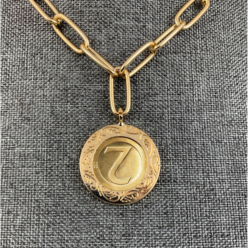 K&ouml;nnte beinhalten: Eine goldene Kettenkette mit einem gro&szlig;en, runden Anh&auml;nger, der den Buchstaben "Z" in einer dekorativen Schriftart zeigt. Der Anh&auml;nger hat ein detailliertes, verziertes Design um den Buchstaben herum.