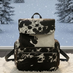 May include: A cowhide backpack with a flap closure and a front zippered pocket. The bag features a black leather handle and straps. The cowhide pattern is black and white, with a soft, textured appearance. The backpack is sitting on a white, fluffy surface.