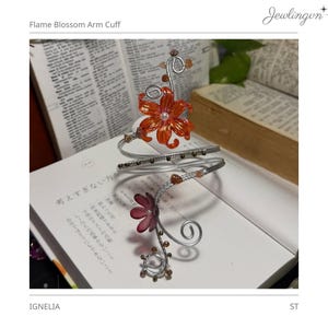 May include: A silver-toned arm cuff with a floral design. The cuff features a large orange flower with a pearl center and smaller purple flowers. The design includes decorative swirls and small, round embellishments. The text "Flame Blossom Arm Cuff" is visible.