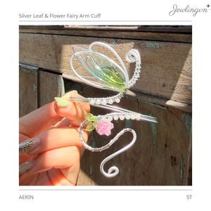May include: Silver fairy arm cuff with leaf and flower details. The cuff is made of silver wire and features a large leaf with green and clear accents, a pink flower, and clear crystals. The text "Silver Leaf & Flower Fairy Arm Cuff" is at the top.