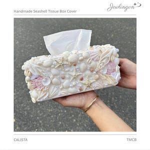 Puede incluir: Funda de caja de pañuelos de conchas marinas hecha a mano. La caja rectangular está adornada con varias conchas marinas, estrellas de mar y perlas en tonos blancos, crema y rosa. Los pañuelos blancos sobresalen de la parte superior. El texto "Handmade Seashell Tissue Box Cover" es visible.