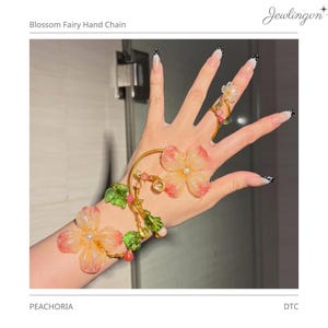 Op de afbeelding: Een Blossom Fairy Hand Chain-sieradenset met een bloemmotief. Het bevat een armband, ring en verbindingsketting, allemaal versierd met doorschijnende roze bloemen, groene bladeren en kleine kralen. De sieraden zijn goudkleurig en bevatten de tekst "PEACHORIA".