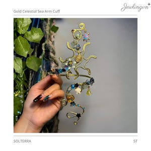 May include: A gold-toned wire arm cuff featuring a sun-shaped design, adorned with various small charms including a turtle, stars, and blue, green, and clear beads. The piece is labeled Gold Celestial Sea Arm Cuff and SOLTERRA at the top and bottom.
