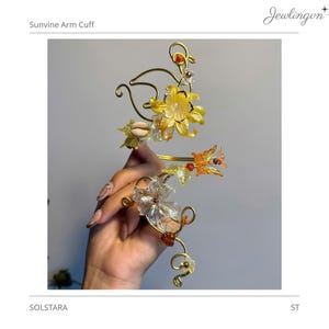 Puede incluir: Un brazalete de alambre dorado con una estructura en forma de enredadera adornada con flores translúcidas amarillas y transparentes, pequeños dijes de mariposa naranja, una concha marina y cuentas rojas y naranjas en los espirales.