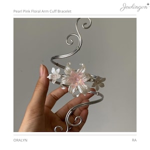 May include: A pearl pink floral arm cuff bracelet. The silver-toned metal cuff features delicate, translucent pink and white flower accents. The bracelet has a curved design with decorative swirls.