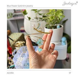 Puede incluir: Joya de brazo dorada con flores azules y detalles de corazón. La joya presenta un diseño delicado con un motivo floral y un dije en forma de corazón. El texto "Blue Flower Gold Arm Jewelry" está en la parte superior de la imagen.